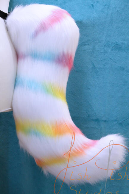A fox styled fursuit tail, mainly in white faux fur with a rainbow spiralised pattern descending from the base to tip. It's design is asymetrical and attaches via belt loops. designed by the commissioner and made by fishpish studios