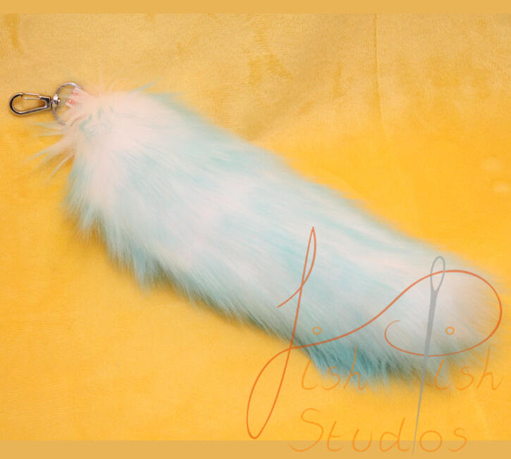 a photo showing a fox styled tail in aqua with a white fade laying on a yellow table. these bag tails are about 9 inches long, and attach to bags or clothing using clips. handmade in the UK by fishpish studios with luxury faux fur