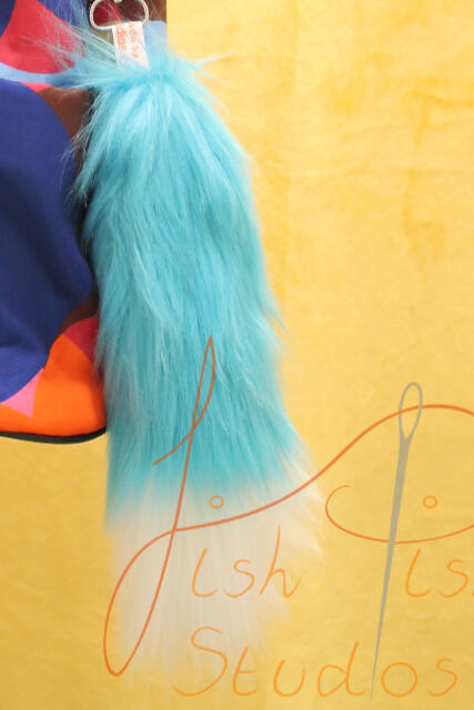 Aqua and White Fox Tail Bag Decoration a photo showing a fox styled tail in aqua with a white tip hanging on a colourful bag. these bag tails are about 9 inches long, and attach to bags or clothing using clips. handmade in the UK by fishpish studios with luxury faux fur
